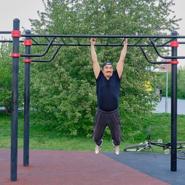 Kıdemli Asyalı adam parktaki çocuk parkında bir çiçek bitkisinin arka planında antrenman yapıyor. Çapraz direkte asılı duruyor. Yaşam tarzı, sağlıklı alışkanlıklar. Hareket bulanıklığı