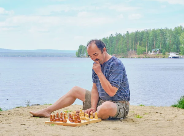 Senior asian man pondering a move in a game of chess and sitting on the beach near the lake. Digital detox concept