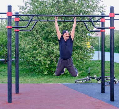 Mutlu kıdemli Asyalı adam şehir parkında bir çiçek bitkisinin arka planında antrenman yapıyor. Çapraz direkte asılı duruyor. Yaşam tarzı, sağlıklı alışkanlıklar. Hareket bulanıklığı