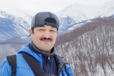 Şapkalı ve mavi ceketli bir turist kameraya bakıyor ve gülümsüyor. Yakın plan. Arkaplan - karla kaplı dağlar.