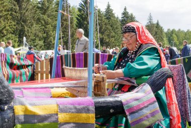 Yekaterinburg, Rusya, 15 Haziran 2019. Bashkir elbiseli yaşlı bir kadın eski bir ahşap odada oturuyor ve halı dokuyor. Şehir parkında ulusal tatil köyü.