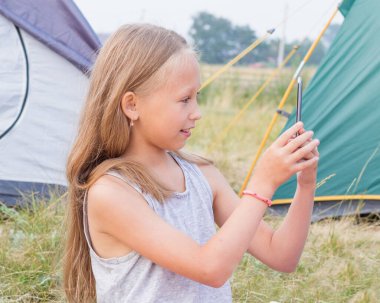 Uzun sarı saçlı şirin küçük kız çadırların arka planında akıllı bir telefonla fotoğraf çekiyor..