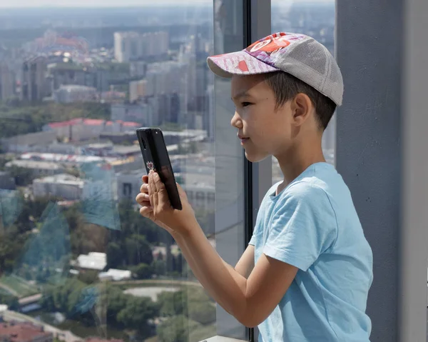 Yekaterinburg, Rusya - 18 Temmuz 2019: Müslüman bir çocuk panoramik bir pencerenin arka planında akıllı telefon kullanıyor. Yüksek bir binadan Yekaterinburg şehrinin bulanık görüntüsü.