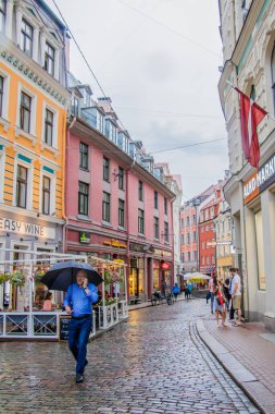 Riga, Letonya - 13 Temmuz 2018: Yağmurlu bir yaz akşamı, turistler yaşlı Riga caddesi boyunca yürür.