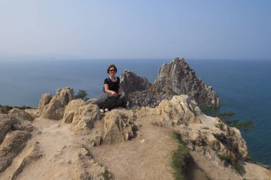 Baykal Gölü. Olkhon Adası. Kıdemli turist kadın kayanın arkasındaki kayanın tepesinde oturuyor..