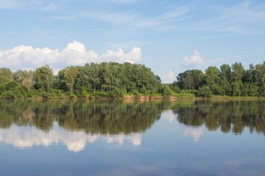 Yaz. Mavi gökyüzü, beyaz bulutlar ve yeşil kıyılar nehre yansıyor. Simetri.