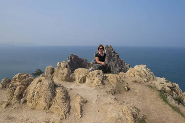 Baykal Gölü. Olkhon Adası. Kıdemli turist kadın kayanın arkasındaki kayanın tepesinde oturuyor..