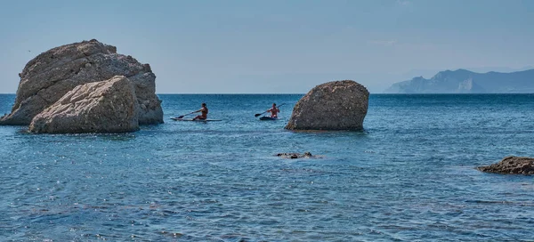 A girl and a guy are floating on sup-surfing among large stones in the sea bay. — Stock Photo, Image