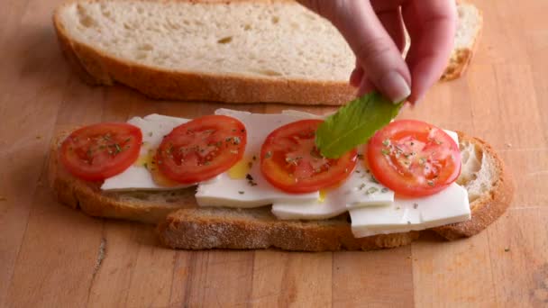 Een vrouw met de hand zet een muntblaadje op een broodje zelfgebakken brood, tomaat, verse kaas en olijfolie — Stockvideo