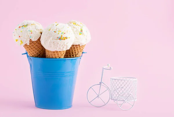 Conos de helado en un cubo azul y una pequeña bicicleta blanca sobre un fondo rosa. Un cono de helado estilo oblea . — Foto de Stock