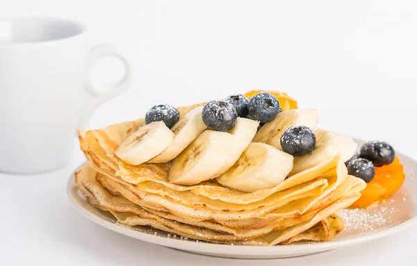En efterrätt på en vit tallrik toppad med frukt och en kopp kaffe. Tunna pannkakor med blåbär och banan — Stockfoto