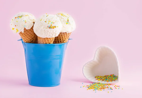 Conos de helado en un cubo azul y un pequeño plato blanco en forma de corazón con cobertura sobre un fondo rosa. Un cono de helado estilo oblea. — Foto de Stock