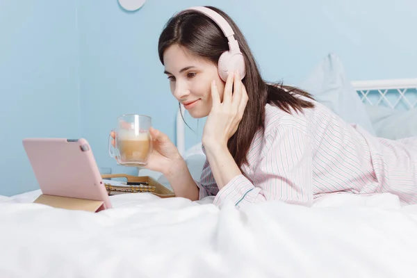 Uśmiechnięta brunetka millenium dziewczyna ze słuchawkami rozmawia z przyjaciółmi przez kamerę internetową, leżąc na łóżku cieszy się rozmową — Zdjęcie stockowe
