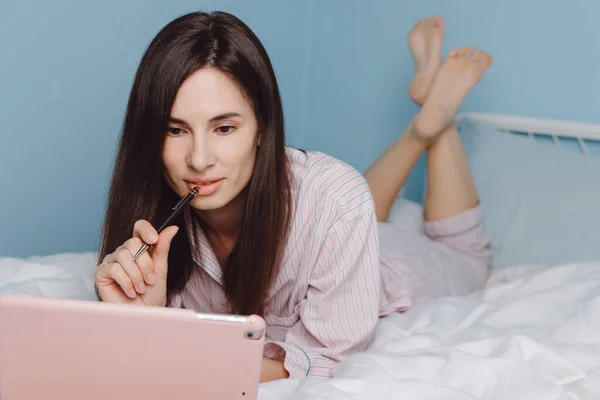 Attractive serious millennial girl lying in bed, making notes, watching webinar or online lecture on digital tablet — Stock Photo, Image
