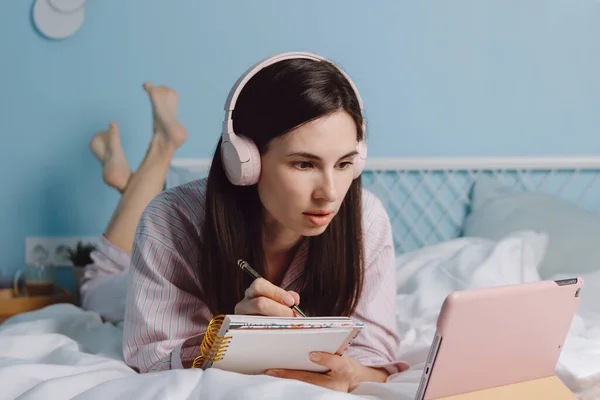 Молода досить спокійна брюнетка тисячолітня жінка в навушниках лежить на ліжку, дивиться вебінар або лекцію за допомогою цифрового планшета — стокове фото