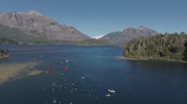 Güneşli bir yaz gününde Nahuel Huapi Gölü 'nü geçen Ironman triatlon yarışmasında yüzücülerin hava görüntüsü. Dağlar, bitki örtüsü ve botlarla çevrili.