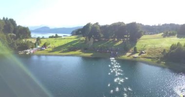 Güneşli bir yaz gününde Nahuel Huapi Gölü 'nü geçen Ironman triatlon yarışmasında yüzücülerin hava görüntüsü. Dağlar, bitki örtüsü ve botlarla çevrili.