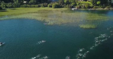 Güneşli bir yaz gününde Nahuel Huapi Gölü 'nü geçen Ironman triatlon yarışmasında yüzücülerin hava görüntüsü. Dağlar, bitki örtüsü ve botlarla çevrili.