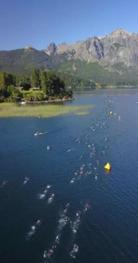 Güneşli bir yaz gününde Nahuel Huapi Gölü 'nü geçen Ironman triatlon yarışmasında yüzücülerin hava görüntüsü. Dağlar, bitki örtüsü ve botlarla çevrili.