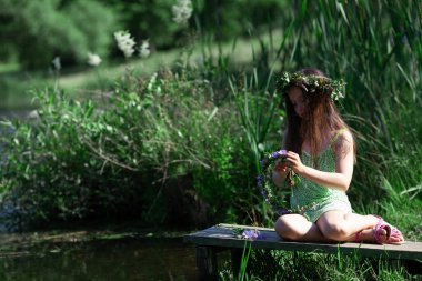 Vahşi çiçeklerin, otların ve yaprakların arasında nehir kenarında yeni çelenkler ören sevimli küçük bir kız. Ivan Kupala 'nın (Aziz John Günü) kutlaması. Seçici odak.