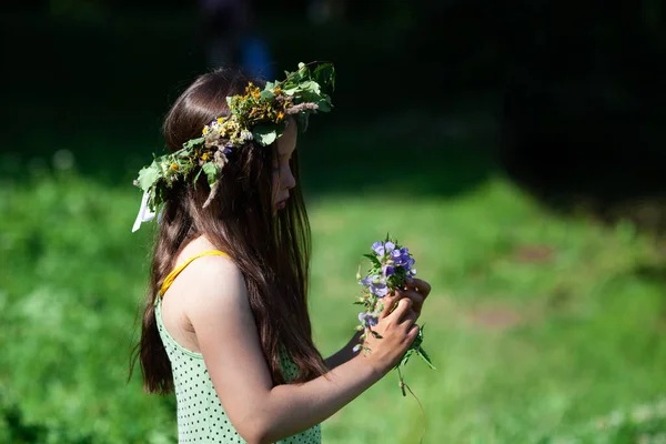 Cute little girl in the wreath of  wild flowers, herbs and leaves weaving new wreath.  Side view. The celebration of Ivan Kupala (St. John Day). Old holiday dedicated to the summer solstice. Selective focus.