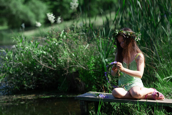 Cute little girl in the wreath of  wild flowers, herbs and leaves weaving new wreath by the river. The celebration of Ivan Kupala (St. John Day). Selective focus.