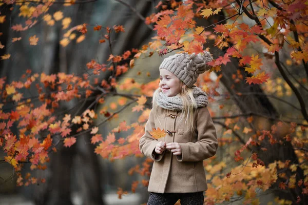 秋の公園で楓の葉を持つ幸せな女の子 可愛いですブロンドの女の子を着てベージュコート ニットキャップとスカーフ笑顔 サイドビュー — ストック写真