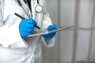 Önlük ve laboratuvar önlüğü giyen ve ameliyat eldiveni giyen bir kadın tıp uzmanı ve bir tablet bilgisayara hasta notları yazarak teknolojiyi kullanıyor.