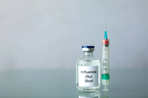 A bottle of a clear medicine influenza flu vaccine along with a syringe and surgical gloves on a plain surface