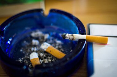 Sigarayı mavi camdan bir kül tablasında yakmak. Sigara tereyağı ve kül yığını, seçici bir odak noktası. 