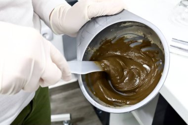 Güzellik salonunda beyaz eldivenli bir kuaför yaşlanma önleyici için deniz yosunlarının kozmetik maskesini karıştırıyor ve plastik bir kabın içindeki cildin plastik çubukla sulandırılması ve cilt tedavisine hazırlanması için.