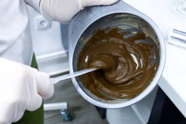 Güzellik salonunda beyaz eldivenli bir kuaför yaşlanma önleyici için deniz yosunlarının kozmetik maskesini karıştırıyor ve plastik bir kabın içindeki cildin plastik çubukla sulandırılması ve cilt tedavisine hazırlanması için.