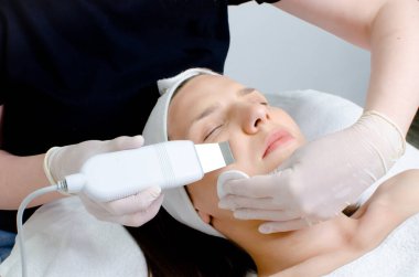 Kozmetik uzmanı, beyaz eldivenli güzellik uzmanı genç bir kadına ultrasonik spatulayla yüz bakımı yapıyor. Ultrasonik spatulayla cilt temizleme tedavisi görüyor. Güzellik salonunda yüz temizleme prosedürü uygulanıyor.