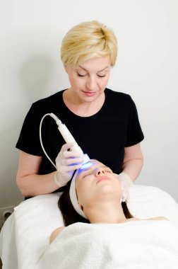 Sarışın güzellik uzmanı, güzellik salonundaki kadın müşteriye mavi ışık terapisi uyguluyor, cilt temizleme için yüz terapisi uyguluyor. Yaşlanma karşıtı tedaviler ve fotoğraf gençleştirme prosedürü, dikey görüntü 