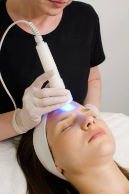 Sarışın güzellik uzmanı, güzellik salonundaki kadın müşteriye mavi ışık terapisi uyguluyor, cilt temizleme için yüz terapisi uyguluyor. Yaşlanma karşıtı tedaviler ve fotoğraf gençleştirme prosedürü, dikey görüntü 