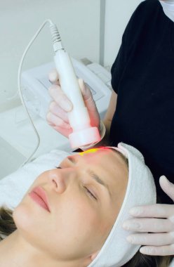 Güzellik uzmanı güzellik salonundaki bayan müşterilere kırmızı ışık terapisi yapıyor cilt temizleme için yüz terapisi yapıyor. Yaşlanma karşıtı tedaviler ve fotoğraf gençleştirme prosedürü, dikey görüntü