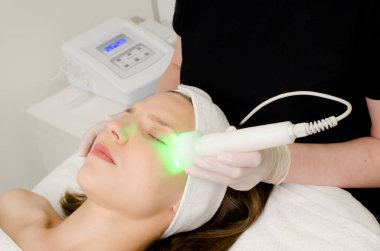 Güzellik uzmanı güzellik salonundaki kadın müşterilere yeşil ışık terapisi yapıyor cilt temizleme için yüz terapisi yapıyor. Yaşlanma karşıtı tedaviler ve fotoğraf gençleştirme prosedürü