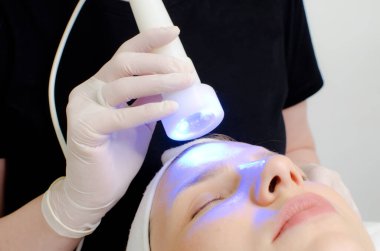 Güzellik uzmanı güzellik salonundaki kadın müşterilere mavi ışık terapisi yapıyor cilt temizleme için yüz terapisi yapıyor. Yaşlanma önleyici tedaviler ve fotoğraf gençleştirme prosedürü, kapatın.