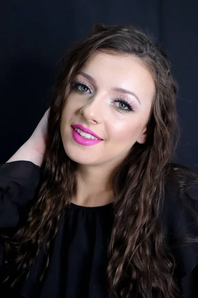 beautiful brunette young girl smiling,looking at camera hand in hair with fresh make up on black background, pink lipstick curly hair over shoulders,in black dress studio shot,vertical image