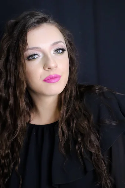 beautiful brunette young girl   looking at camera with fresh make up on black background, pink lipstick curly hair over shoulders, in black dress  studio shot, vertical image