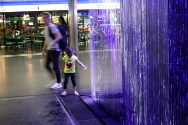 Zürih, İsviçre, 19 Temmuz 2019: Tren istasyonunun yeraltı geçidinde bulunan renkli çeşmeye yaklaş. Zürih ana tren istasyonunun içi, yolcular aceleyle geçerken.