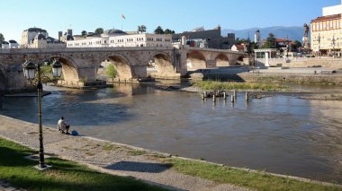 Üsküp, Kuzey Makedonya, 27 Haziran 2019: Üsküp şehir merkezindeki Vardar Nehri üzerindeki taş köprü yakınlarındaki balıkçı yaz günü mavi gökyüzü ile Makedonya Arkeoloji Müzesi yakınlarında