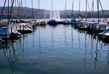Zürih, İsviçre, 23 Temmuz 2019: güneşli bir günde suyun yansıması olarak Zürih Gölü 'ndeki çeşmenin yanındaki rıhtımdaki park halindeki tekneleri kapladı.