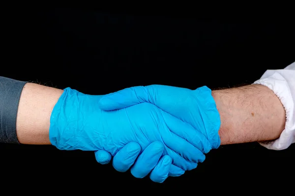 Shaking hands, two people wearing blue latex gloves shake hands. Only hands visible. Closeup view, black background.