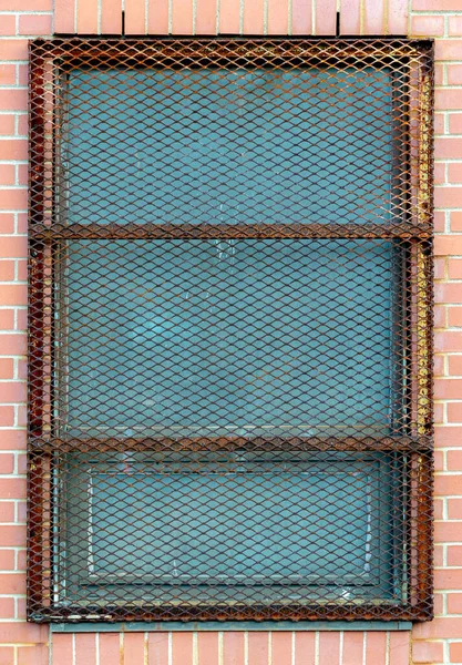 A heavy metal screen protecting the window in a brick building. The screen has a diamond shape pattern and is rusty.
