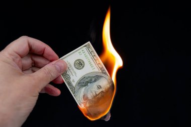 100 dolarlık banknotu yakmak. Bir el yanan banknotu tutuyor, yaklaşık yarısı yanmış. Siyah arkaplan.
