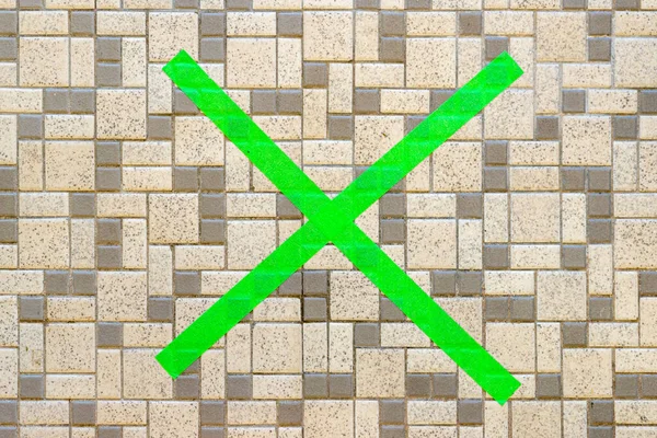 A green social distancing X on a tile floor, made from masking tape. Top view looking directly down, closeup view.