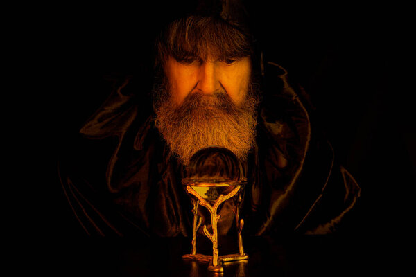 Wizard gazing into a crystal ball. He wears a dark cloak. Shallow depth of field, focus on face. Dark scene illuminated by light from fire.