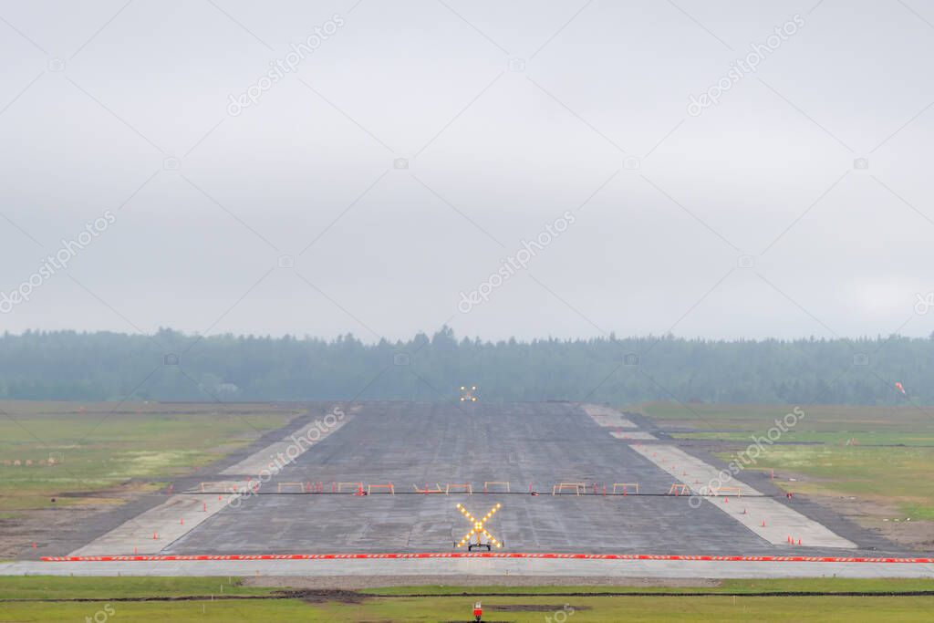 Closed runway at an airport on an overcast, foggy day. The runway has a large illuminated warning X sign at each end, and is blocked by sawhorses.