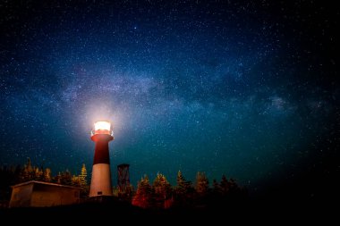 A lighthouse at night with a star filled sky above. The light in the top of the lighthouse is illuminated. The Milky Way is visible. clipart